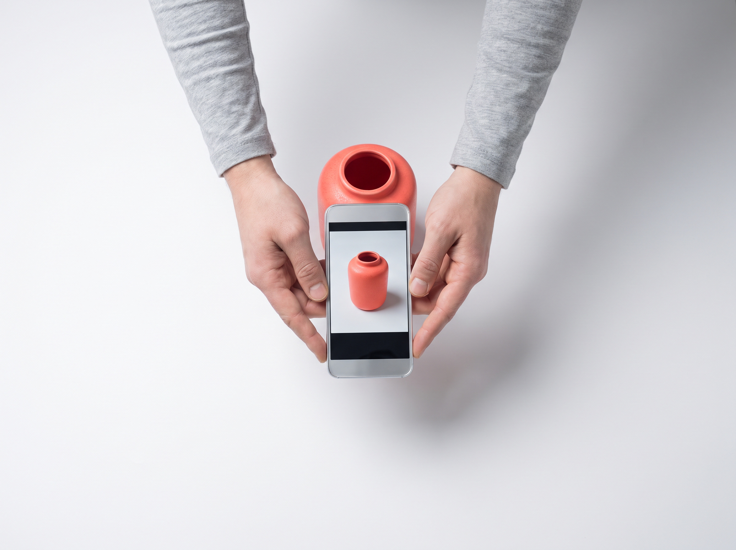 Hands photographing a coral vase on a white sweep with a phone